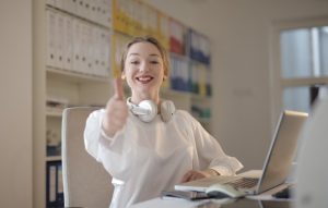 Girl in all white with white headphones giving thumbs up while working on a laptop