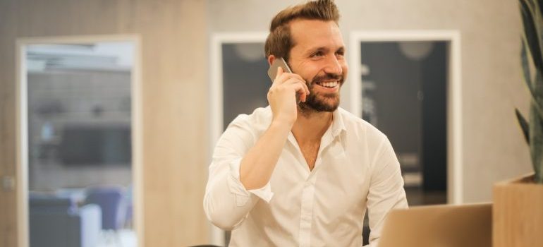 A man talking on the phone next to a laptop and a cup of coffee.