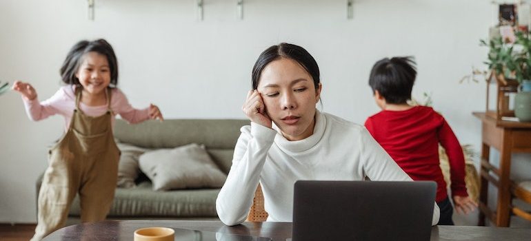 Woman sitting at the table and working on computer and 2 children running behind her