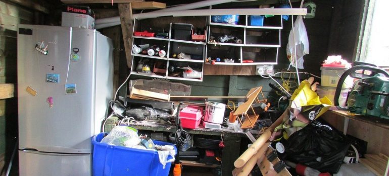 a messy room cluttered with various things, an overloaded shelf on the wall and a refrigerator with magnetic pictures