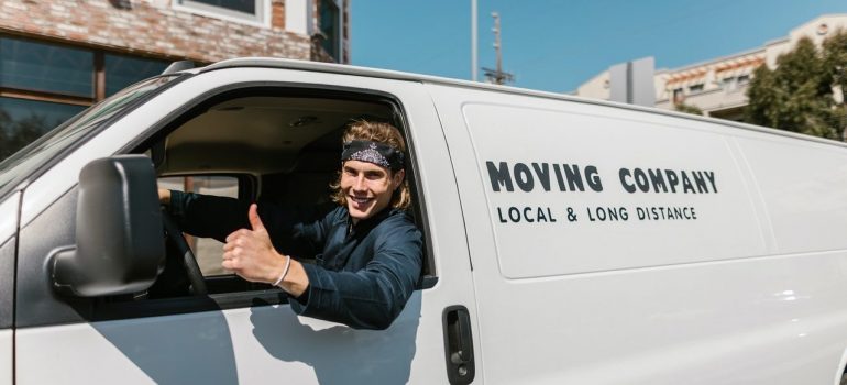 Man in black clothes seating in a white van with inscription has timely arrived to make your movers cost per hour lower and is in front of the house 