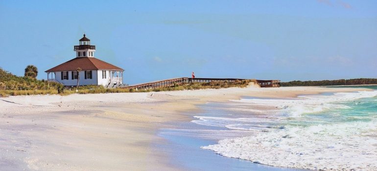 Sandy beach, waves on the water and in the distance a lighthouse and a person walking over the pier.