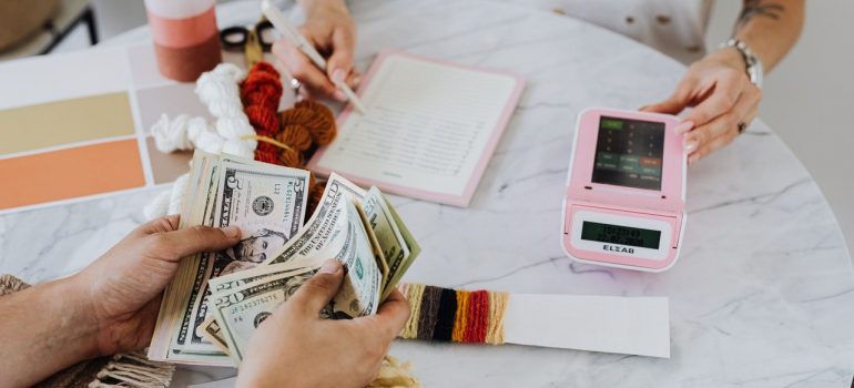 Man’s hands counting money and the woman hands are checking the list and using the calculator which lie on the white marble slab of the table. 