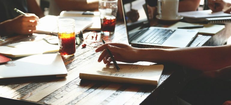 A table full of papers and a laptop, two glasses and a cup on it, and the two men at front holding pens.