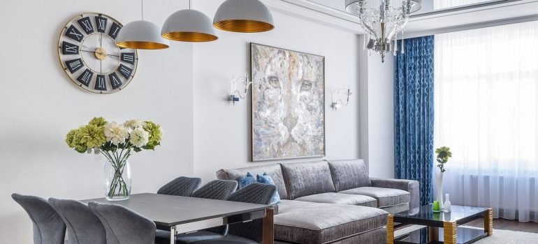 A living room combined with dinning table, chairs, modern sofa, picture and clock on the wall, chandeliers, carpet, and floor to ceiling window.