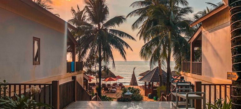 Two houses and a path walk between them, leading down to the beach with palms, parasols and the ocean in the distance. 