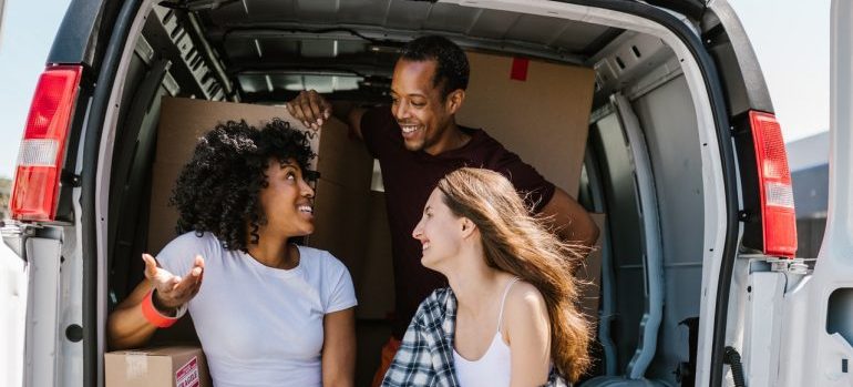 A group of three people sitting in the back of a van with boxes behind them, talking