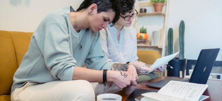 Two woman reading papers seating on sofa, with a coffee table in front, with a laptop, notebook and coffee on it. 