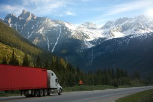 A red moving truck on the road with green hills and mountains covered with snow, driving from Miami to Los Angeles. 