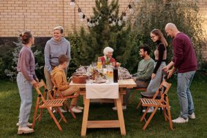 A group of people are sitting and standing around a table in the yard, where food for the party has been arranged.