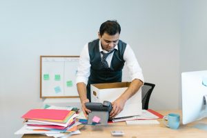 A man in office is collecting stationary from the table and putting it into a cardboard box. 