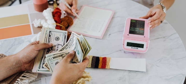 People sitting around the table, holding dollar bills and using calculator, pen and notebook, checking funds for moving to Miami on a budget.