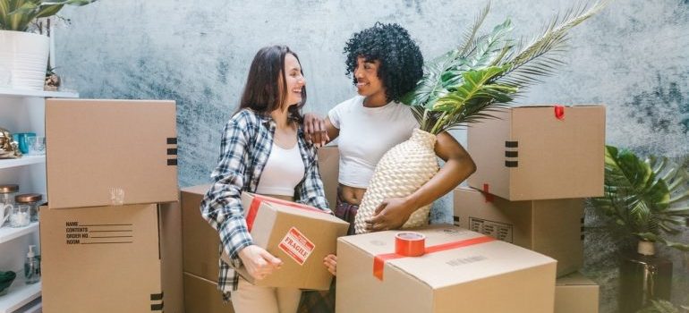 two women surrounded by boxes while one is hodlinga box and teh other a vase with flowers